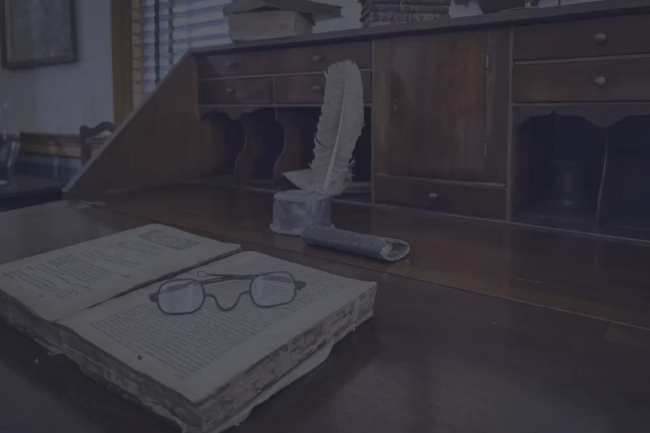 Desk with a folded newspaper and a pair of reading glasses resting on dark wood, a feather quill in a holder nearby.