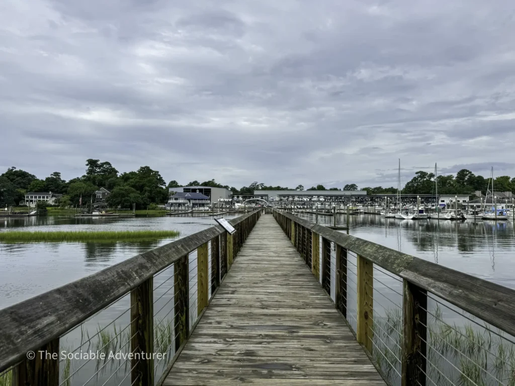 Wooden pier extending toward a marina with sailboats docked along calm water under a cloudy sky.