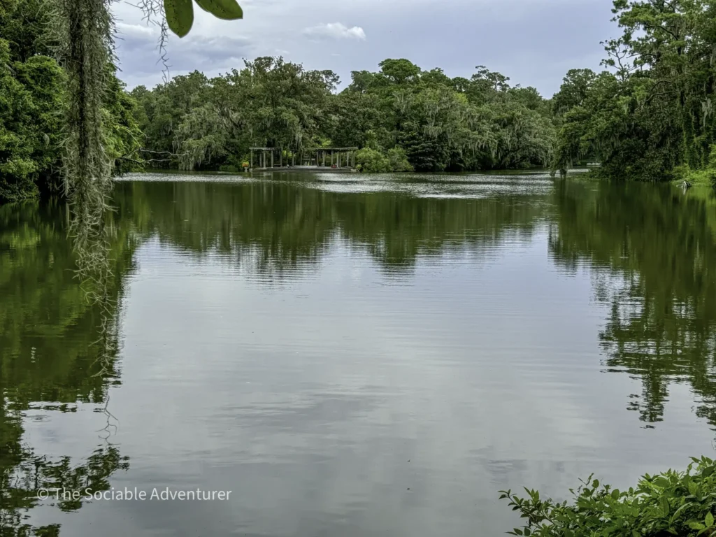 Tranquil lake surrounded by dense green trees with Spanish moss hanging from branches and a small pergola visible on the far shore.