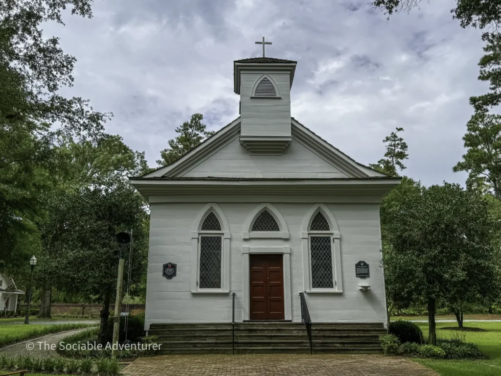 White church with a red door, steeple, cross on top, and arched windows surrounded by trees and greenery.