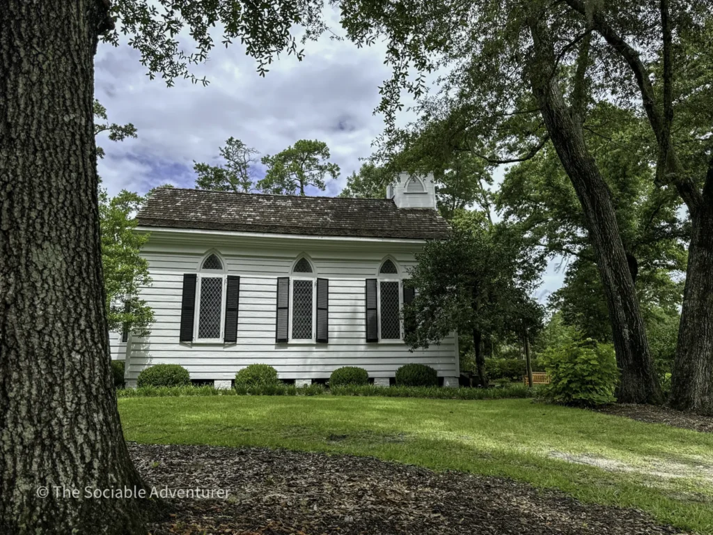 White clapboard house with arched windows and black shutters, framed by tall trees and a green lawn in the foreground.
