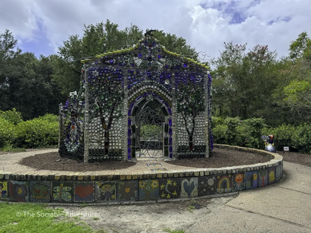 Colorful bottle-cap mosaic archway in a garden, made from reused glass bottles, sunlit.