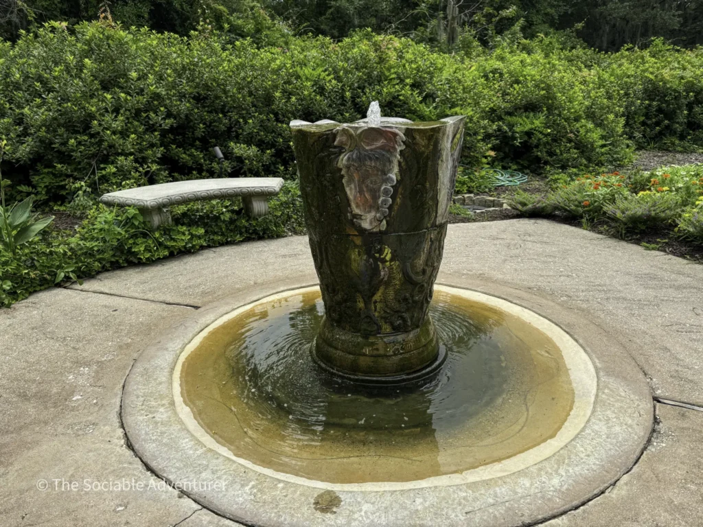 Stone garden fountain: a carved, vase-shaped sculpture with water spouting from the top, set in a circular basin amid greenery and a concrete path.