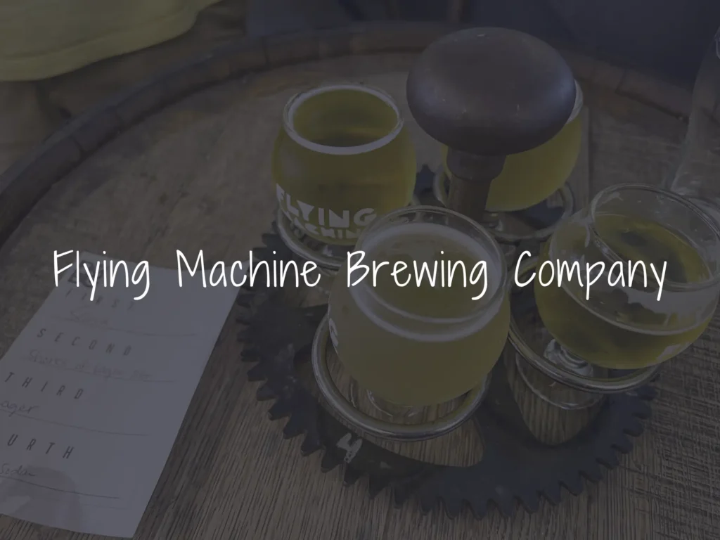 Close-up of a beer brewing setup on a wooden table with small glasses of beer and a yellow can; overlaid text reads 'Flying Machine Brewing Company'.